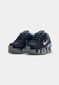Melna un pelēka Nike Shox skrienšanas apavu pāris ar gaiši ziliem akcentiem un mīkstinātām atsperēm uz balta fona.