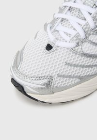 Zapatilla de correr de malla blanca con detalles en plata, con parte superior texturizada, cierre de cordones y suela de goma con detalles negros.
