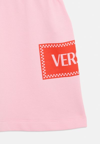 T-shirt de algodão rosa com um logótipo retangular impresso em vermelho e texto branco "VERS." e uma borda quadriculada perto da bainha. Textura suave.