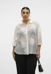 Weiße bestickte Bluse mit Dreiviertelärmeln, Knopfverschluss und transparenter Textur. Kombiniert mit schwarzen Hosen und einer kleinen schwarzen Tasche.