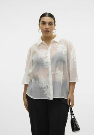 Blusa bianca ricamata con maniche a tre quarti, chiusura a bottoni e texture trasparente. Abbinata a pantaloni neri e tiene una piccola borsa nera.