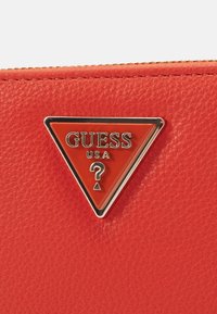 Portefeuille en cuir rouge texturé avec un emblème métallique triangulaire portant l'inscription "GUESS USA" et un point d'interrogation, ajoutant un détail de design distinctif.