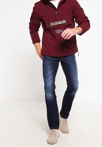Bordeaux pullover met een halve rits, grote patch en Scandinavische vlag; gecombineerd met donkerblauwe spijkerbroek en lichtbeige schoenen.