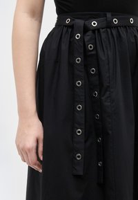 Αριστερό χέρι και μπράτσο ενός ατόμου που φοράει μια μαύρη φούστα με πιέτες και μια μακριά ζώνη με ασημένιες τρύπες.