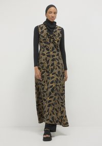 Maxikleid in Schwarz mit goldenen Blattmustern, mit einem Bindedetail am Halsauschnitt. Getragen über einem schwarzen Langarmshirt, kombiniert mit schwarzen Sandalen.
