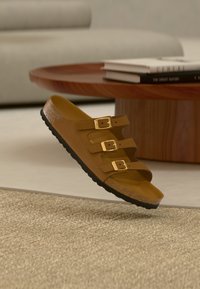 Καφέ σανδάλι με τρεις χρυσές πόρπες, σόλα από φελλό και μαύρη σόλα, αιωρούμενο πάνω από χαλί μπεζ με υφή σε σαλόνι.