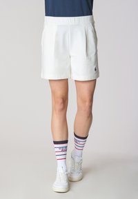 Pantaloni corti bianchi su misura con pieghe frontali, abbinati a sneakers bianche e calze a righe blu navy, rosse e bianche. Tessuto morbido e design strutturato.