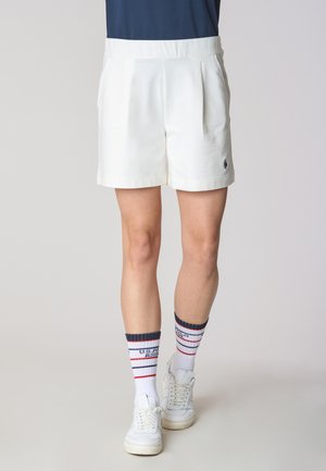 Pantaloni corti bianchi su misura con pieghe frontali, abbinati a sneakers bianche e calze a righe blu navy, rosse e bianche. Tessuto morbido e design strutturato.