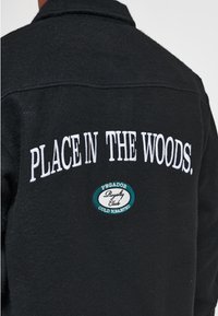 Chaqueta de forro polar negra con texto bordado en blanco "PLACE IN THE WOODS." Incluye un parche redondo con detalles en verde azulado y elementos de marca.