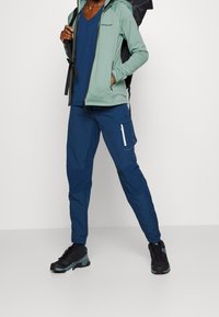 Veste zippée vert clair, chemise bleue à col en V, pantalon cargo bleu marine avec poche zippée, chaussures noires avec accents gris, sur un fond neutre.
