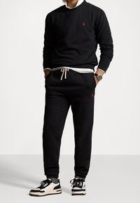 Svart sweatshirt med en röd logotyp, kombinerad med svarta sweatpants med fickor och en krämfärgad dragsko. Bäres med svartvita sneakers.