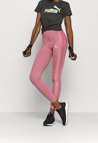 Sportowy strój Puma składający się z czarnego T-shirtu z nadrukiem w wężowy wzór i złotym logo, różowych legginsów z białym emblematem oraz czarnych butów sportowych.