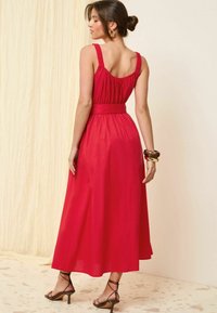 Rochie maxi roșie cu un corset ajustat, bretele elastice și talie cu curea. Țesătură fluidă și textură netedă. Pantofi cu toc negri și curea.