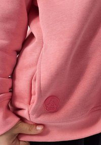 Felpa rosa con tessuto liscio e polsini a coste. Presenta una targhetta con il logo JOTT sul lato e una tasca a marsupio.