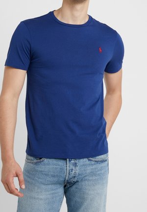 Homme portant un t-shirt bleu uni à manches courtes avec un petit logo rouge sur le côté gauche de la poitrine, associé à un jean bleu clair.