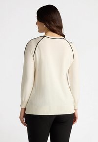 Hellbeiger, gestrickter Pullover mit geripptem Saum und Bündchen, der einen schwarzen Akzent entlang der Schultern aufweist. Glatte Textur mit subtiler Schimmer.