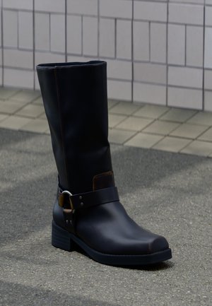 Zwarte leren laars tot halverwege de kuit met een lage hak en een decoratief riempje met metalen gesp, staand op een grijs bestraatte ondergrond nabij een betegelde muur.