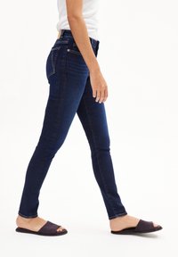 Jeans skinny bleu marine avec des détails de couture subtils, taille haute et poches arrière, associés à des sandales à glissière noires.
