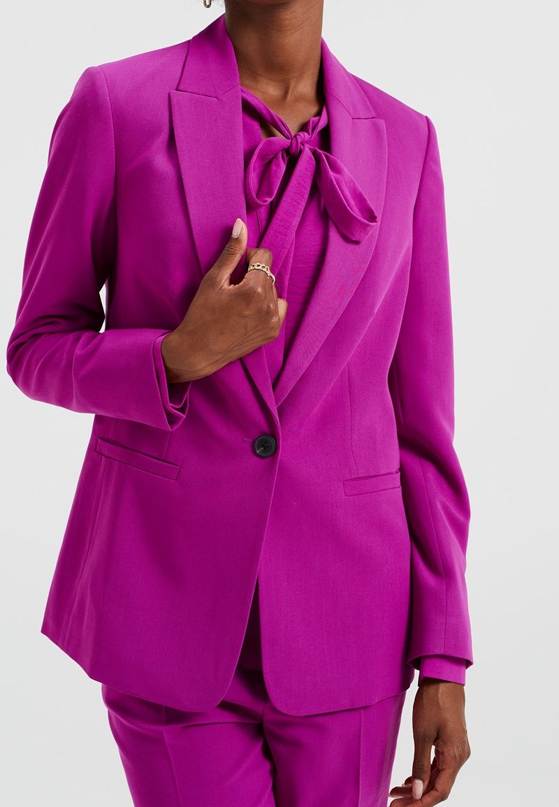 Fuchsia pantsuit featuring a tailored blazer with notched lapels and a matching blouse with a tied neck detail. Smooth fabric, sleek design.