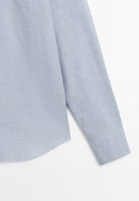 Manche et côté d'une chemise bleu clair à manches longues en tissu subtilement texturé sur fond blanc.