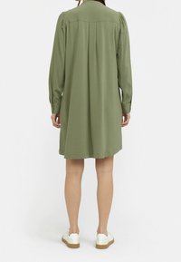 Oliwkowa sukienka z długimi rękawami, zapinana na guziki z tyłu, z plisowanymi detalami i luźnym krojem, w zestawieniu z białymi trampkami.