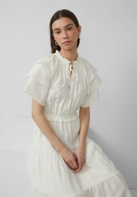 Witte jurk met geschulpte korte mouwen, een koordsluiting bij de hals en een elastische taille. De stof heeft een zachte, lichte textuur met geplooide lagen.