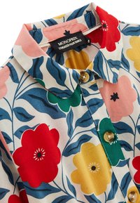 Chemise à boutons avec un motif floral rouge, jaune et rose sur fond blanc, col montant et boutons beiges.