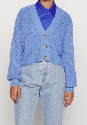 Lichtblauwe gebreide cardigan met V-hals, drie grote knopen en geribbelde manchetten. Gecombineerd met een blauwe overhemd met kraag en lichte spijkerbroek.