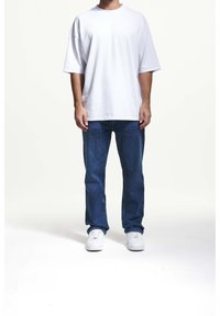 Biała, bawełniana, oversize'owa koszulka z krótkim rękawem, zestawiona z niebieskimi dżinsami i białymi trampkami, stoi na jednolitym tle.