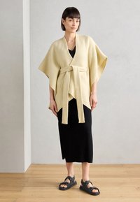 Beige, teksturoitu takki soljella, mustan ribbimekon ja mustien sandaalien päällä. Ominaisuuksiin kuuluu leveät hihat ja suora helma.