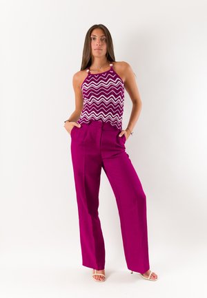 Giovane donna in piedi con le mani nelle tasche, che indossa un top smanicato viola e bianco a zigzag, pantaloni magenta a vita alta e sandali beige.