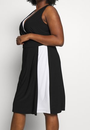 Femme portant une robe noire sans manches avec de larges panneaux verticaux blancs, debout devant un fond uni.