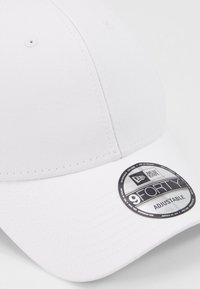 Weißes Baseballcap aus Baumwolle mit gebogenem Schirm und verstellbarem Rücken. Enthält ein rundes Etikett mit der Aufschrift "9FORTY ADJUSTABLE."