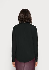 Person mit schulterlangem, welligem, dunklem Haar, die ein schwarzes Langarmshirt und einen dunkelpurpurfarbenen Rock trägt und sich von der Kamera abwendet, vor einem schlichten Hintergrund.