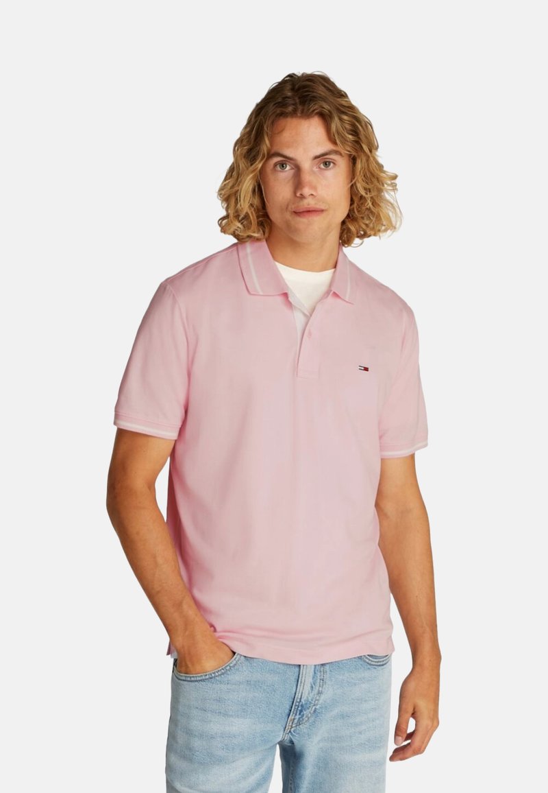Camisa polo de color rosa claro con cuello clásico y mangas cortas, que presenta un pequeño logo bordado en el pecho. Se lleva con unos jeans azul claro.