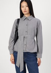 Grå skjorta med krage, långa ärmar, insnörd midja och en slät textur. Matchad med blå denimjeans och en svart väska.