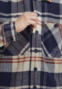 Flanelinė marška su tamsiai mėlyna, kremine ir oranžine languota raštą. Savybės: mygtukų užsegimas, dvi krūtinės kišenės ir švelni vilnai panaši tekstūra.