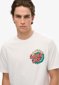 Camiseta de algodón blanca con un gráfico colorido en estilo cartoon de "Super Dry" con un diseño de ola en el área del lado izquierdo del pecho.