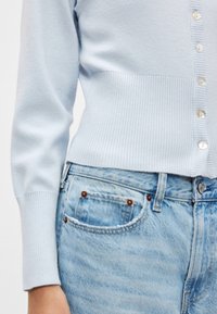 Ανοιχτό μπλε πλεκτή ζακέτα με ριμπ τελείωμα και μανσέτες, διακοσμημένη με κουμπιά. Συνδυασμένη με ξεθωριασμένο μπλε τζιν παντελόνι.