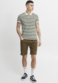 Tricou polo cu dungi în verde și crem, cu guler și trei nasturi. Asortat cu pantaloni scurți verde măsliniu și adidași negri.