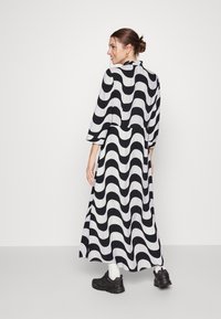 Vestido maxi preto e branco com padrão, apresentando mangas fluídas e um design ondulado. O modelo usa ténis pretos e meias brancas.