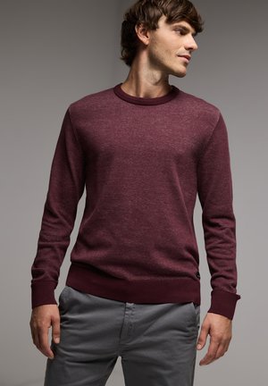 Bordeaux gebreide trui met een ronde halslijn en lange mouwen; ribgebreide manchetten en zoom; gladde textuur; gedragen met grijze broek.