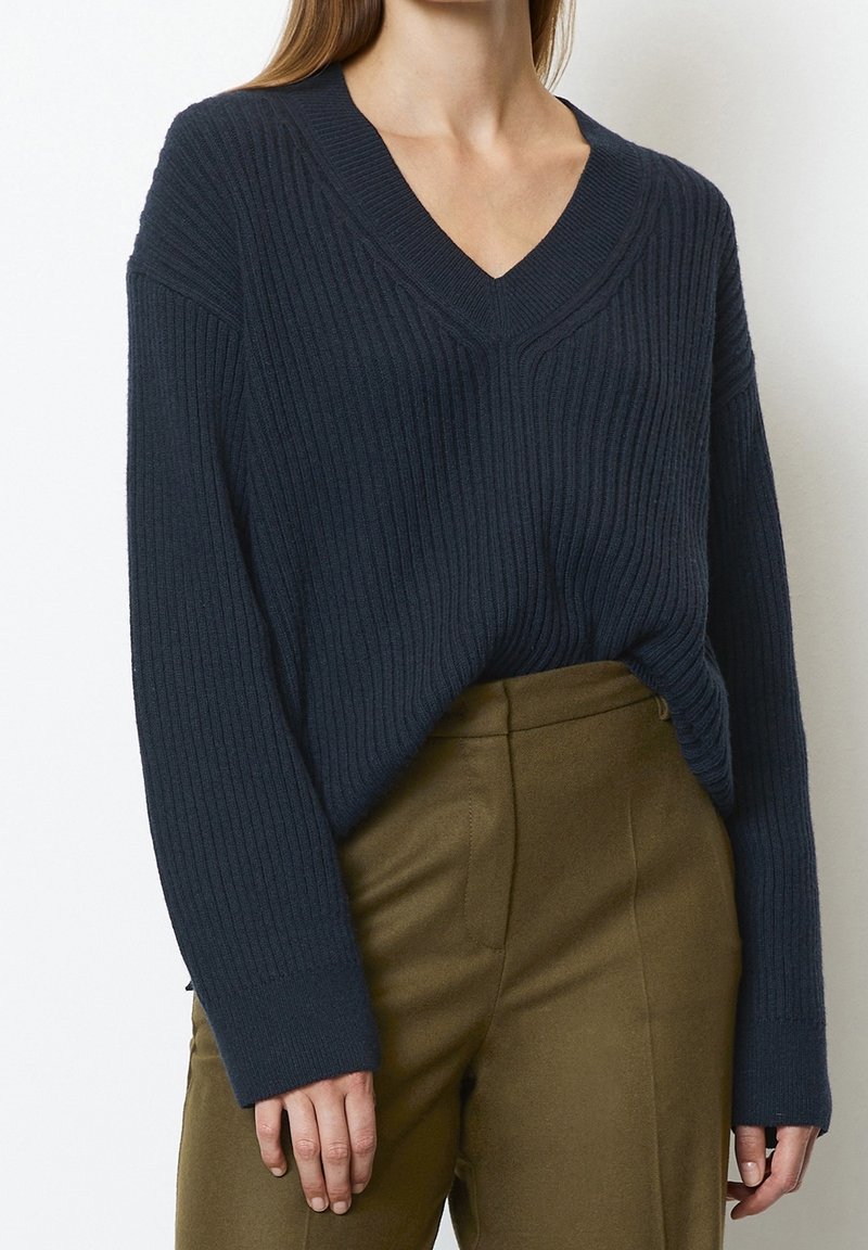 Pull en maille côtelée bleu marine avec col en V et manches longues, présentant une coupe décontractée et un tissu texturé, associé à un pantalon tailleur vert olive.