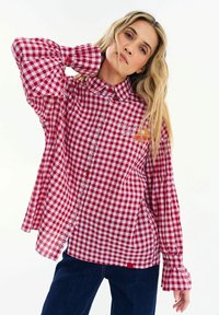 Žena s dlouhými blond vlasy, oblečená v červenobílém košili s nabíranými rukávy, pózující s jednou rukou za krkem.