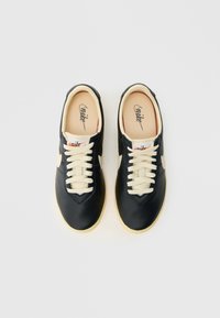 Mustad nahast tossud kreemikaspaeltega, beežide talladega ja logoga keelel. Ümar nina ja sile tekstuur, millel on õmblusdetailid.