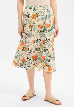 Femme portant une jupe beige mi-longue à motifs floraux orange, vert et bleu et des sandales plates beiges à enfiler.