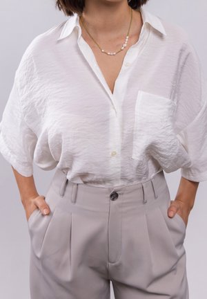Femme portant une chemise blanche ample boutonnée, un pantalon gris taille haute et un collier en or avec des perles blanches, les mains dans les poches.