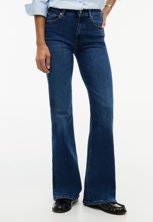 Person trägt dunkelblaue ausgestellte Jeans, hellblaues Hemd mit hochgekrempelten Ärmeln, schwarze Schuhe und eine goldene Armbanduhr auf weißem Hintergrund.