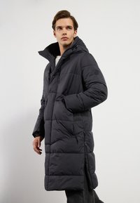 Doudoune gris foncé avec capuche, présentant un design matelassé, des poches zippées et une silhouette longue, fabriquée en matériau isolant.