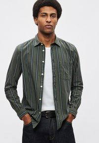 Koszula na guziki w oliwkowym odcieniu z białymi i granatowymi paskami, z kieszenią na piersi i dopasowanymi mankietami, noszona na białym t-shircie.